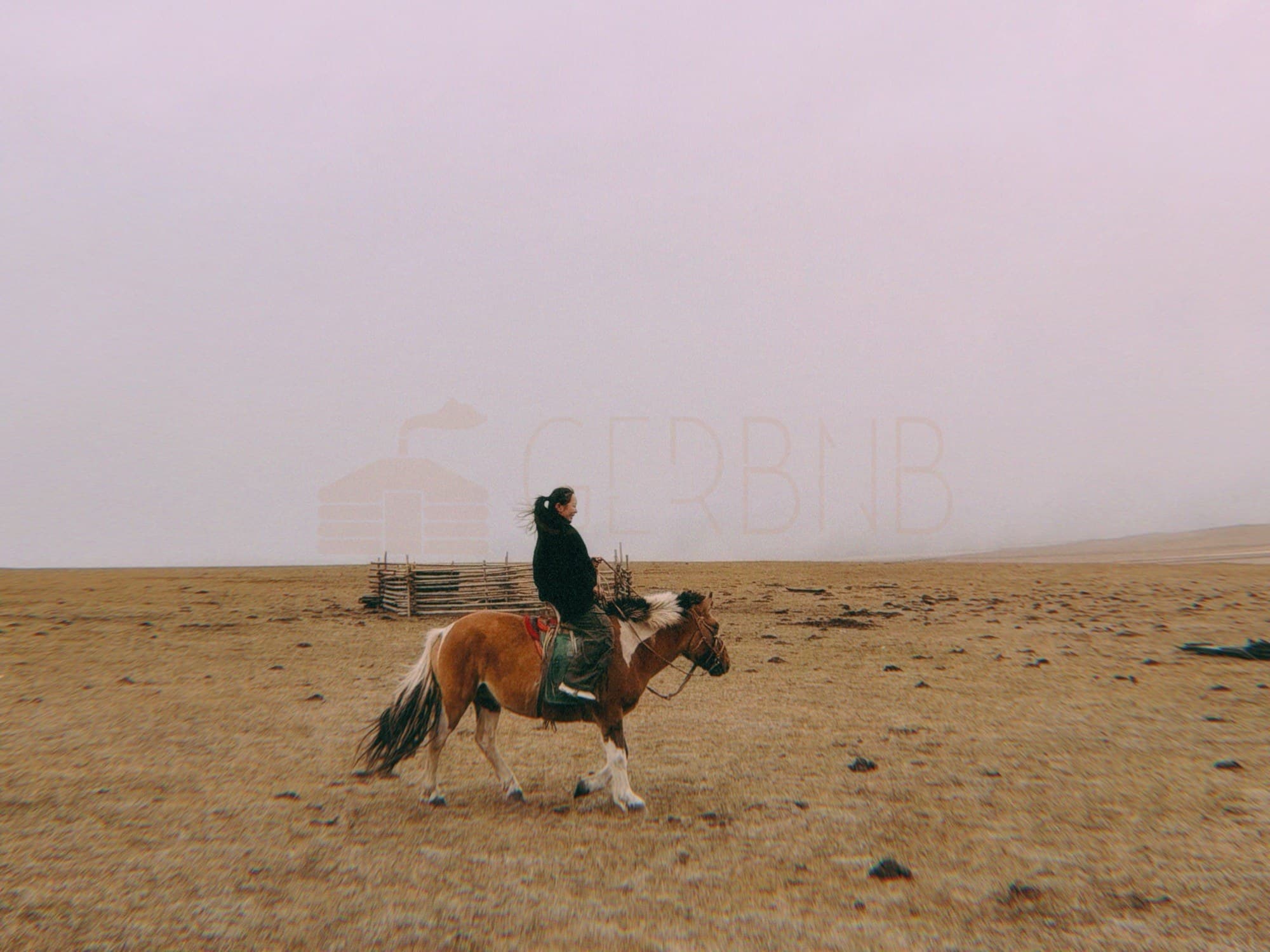 A rider on a pinto horse crossing foggy steppe pasture, a wooden corral behind.
